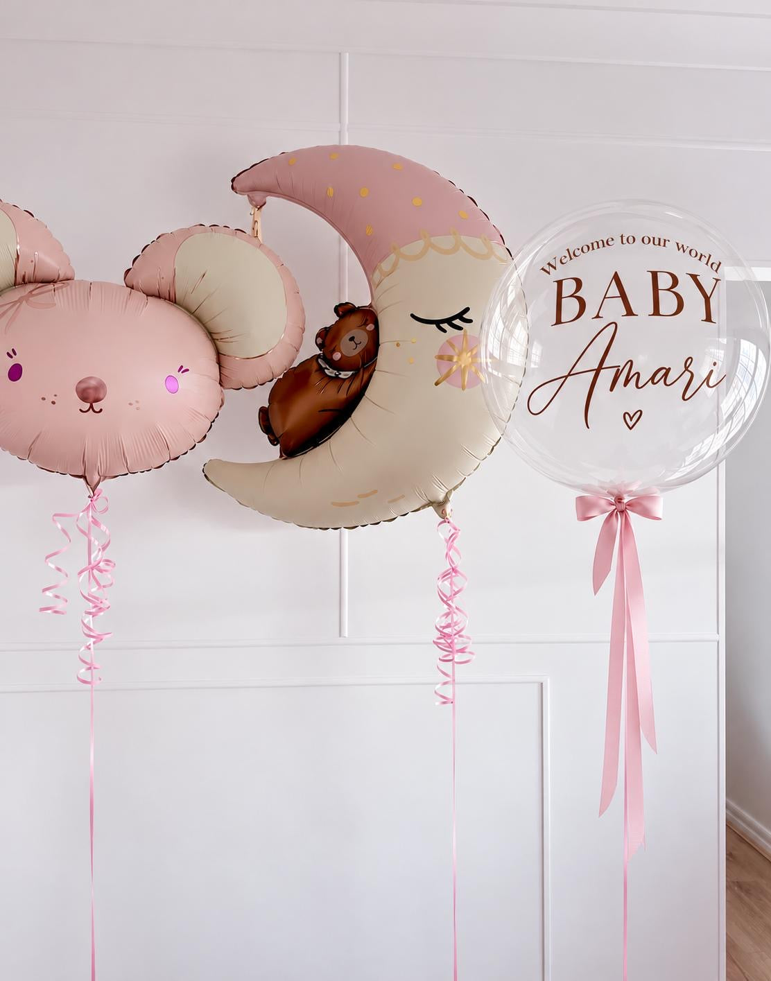 Pink and beige baby shower balloons including a bear head, crescent moon with a teddy bear, and a clear round balloon that reads 'Welcome to our world BABY Amari' with a pink ribbon, set against a white paneled wall.