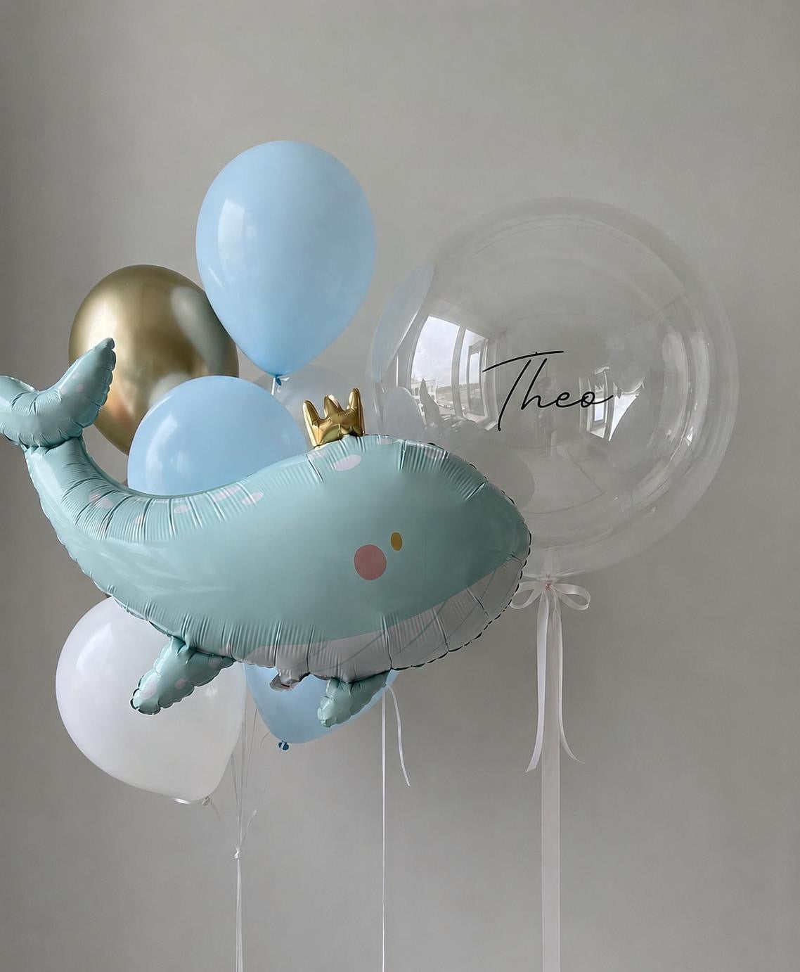 Personalised whale fish balloon bouquet with pastel blue, white and gold helium balloons on a neutral background.