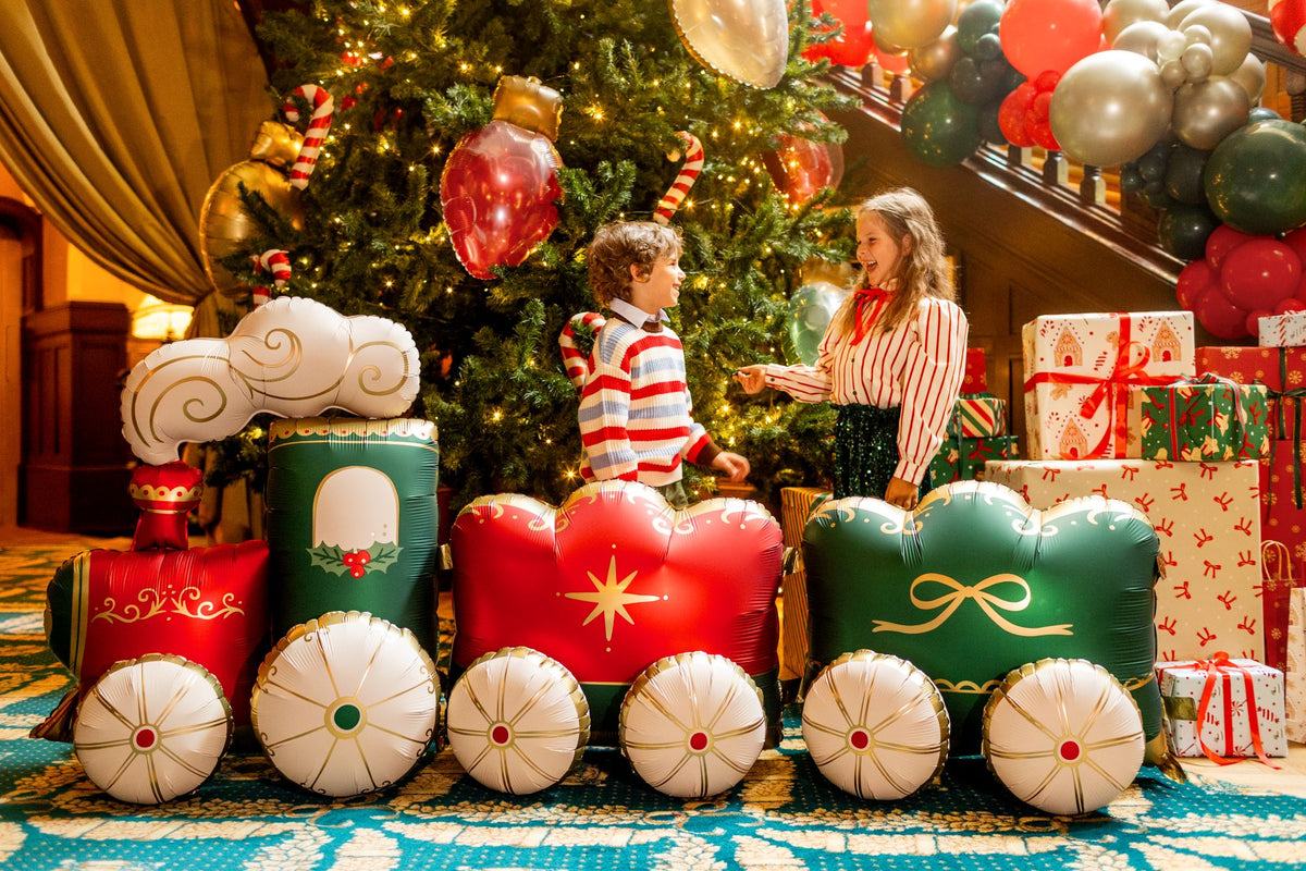 The Christmas Express Edit XXL Christmas train balloon displayed beside a decorated Christmas tree, featuring festive red and green carriages with elegant gold detailing.